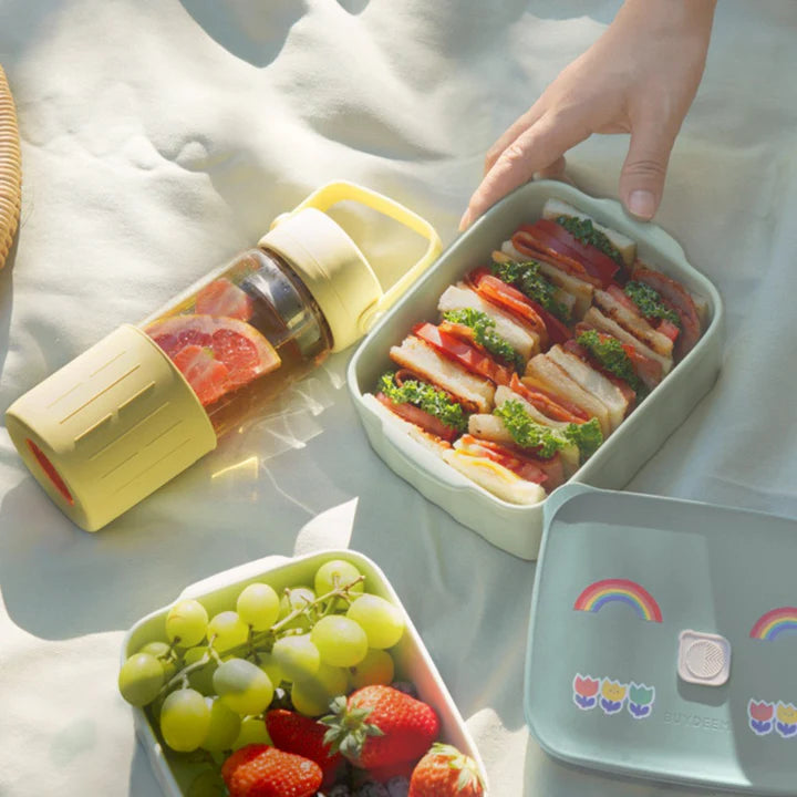 Bento box with sandwiches, fruits, and a bottle on a light surface.